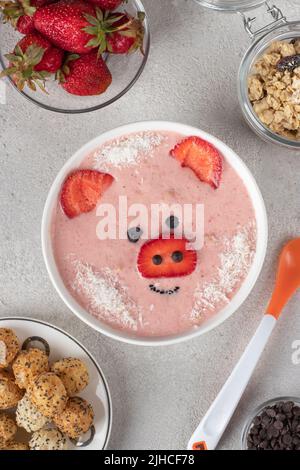 Divertimento per i bambini - ciotola di frullato in forma di maialino divertente, con fragole, granola, cioccolato e palle croccanti, vista dall'alto su sfondo grigio Foto Stock