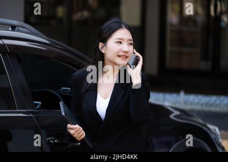 Gli uomini d'affari asiatici, i proprietari di affari, i presidenti di azienda o i dipendenti femminili che parlano al telefono e che tengono un tablet stanno uscendo dall'automobile a. Foto Stock