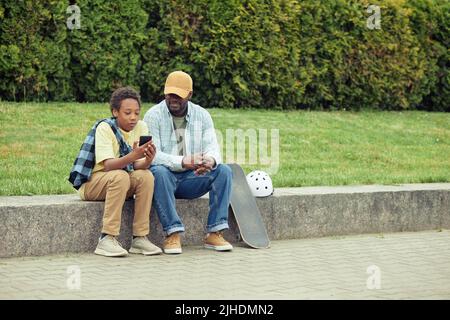 Ragazzo che mostra le foto sul telefono cellulare al papà mentre riposano all'aperto dopo lo skateboard Foto Stock