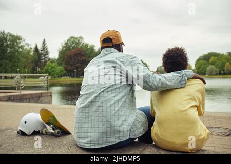 Vista posteriore del papà che abbraccia suo figlio e si riposano insieme dopo lo skateboard guardando la bella vista del fiume Foto Stock