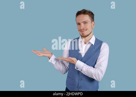 Giovane uomo d'affari in camicia bianca e abito blu gilet isolato su sfondo blu Studios ritratto. Concetto di affari di ricchezza di successo di carriera. Mostra un gesto verso una copia del luogo e sorride Foto Stock