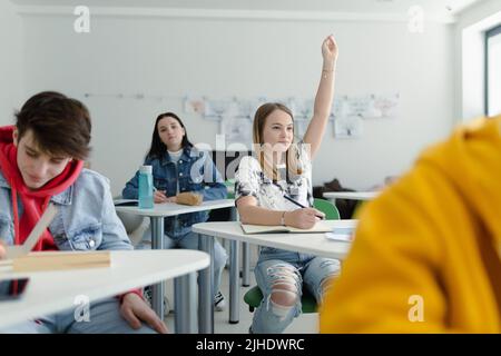 Gli studenti delle scuole superiori prestano attenzione in classe, seduti sulle loro scrivanie e alzando la mano. Foto Stock