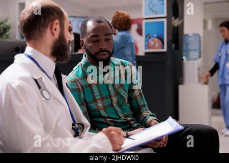 Medico che parla con il paziente di sesso maschile in sala d'attesa, utilizzando il rapporto di controllo medico per dare la diagnosi e il trattamento della malattia. Medico e uomo che fa la consultazione sanitaria in clinica. Foto Stock