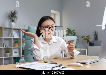 Frode. Sconvolto giovane bella donna asiatica in possesso di una carta di credito. Una giovane donna delusa ha perso il suo risparmio di vita a una truffa. Seduti alla scrivania in ufficio Foto Stock