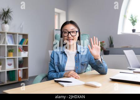 Apprendimento a distanza. Una giovane donna asiatica è un insegnante, conduce lezioni online, lezioni. Guarda nella fotocamera, spiega, dice. Seduti in bicchieri e jeans a un tavolo di un computer in un'aula Foto Stock