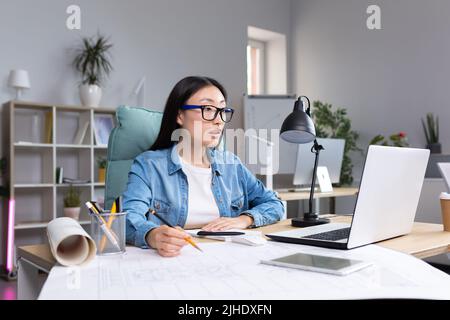 Giovane ragazza asiatica bella architetto designer indossare occhiali che lavorano al computer desktop e disegnare con matita, lavorando al progetto. Foto Stock