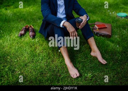 Uomo d'affari con caffè, riposo e seduta a piedi nudi nel parco, sentirsi liberi, fuggire dal lavoro, concetto di equilibrio della vita lavorativa. Foto Stock