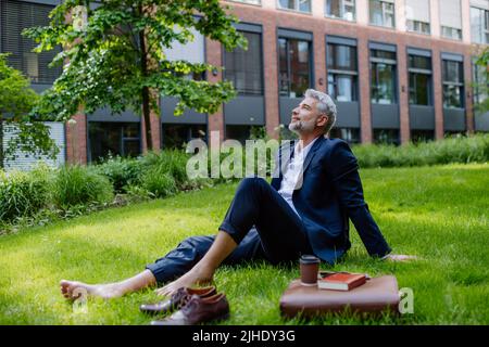 Uomo d'affari maturo che riposa e siede a piedi nudi nel parco, sentendosi libero, fuggendo dal lavoro, concetto di equilibrio di vita di lavoro. Foto Stock