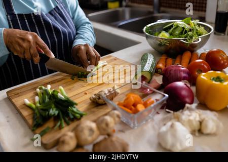Sezione centrale di donna matura biraciale che indossa grembiule tritare verdure sul tagliere in cucina Foto Stock