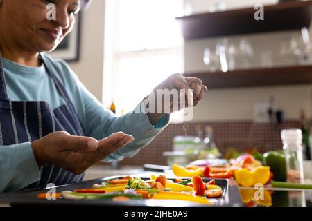 Metà sezione di donna matura biraciale indossando grembiule cospargere spezie su verdure in vassoio in cucina Foto Stock