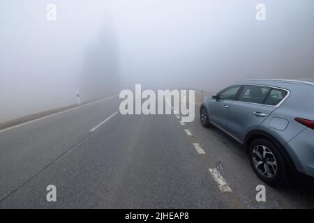 Un'auto SUV grigia è parcheggiata sul lato di una strada vuota e rettilinea invasa da una fitta nebbia. Sullo sfondo, si possono indovinare le sagome di alcuni alberi alti, avvolti dalla nebbia. Foto di alta qualità Foto Stock