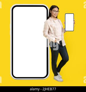Mostrando schermo bianco sul telefono affascinante metà età asian donna appoggiata indietro su smartphone gigante, enorme con schermo bianco indossando casual isolato su sfondo giallo. Spazio libero mock up. Foto Stock
