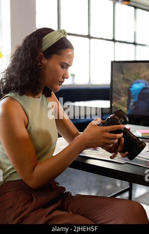 Giovane donna d'affari biraciale che esamina la macchina fotografica alla scrivania in ufficio creativo Foto Stock