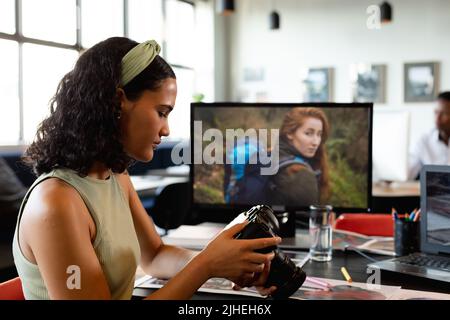 Giovane donna d'affari biraciale che esamina la macchina fotografica alla scrivania in ufficio Foto Stock