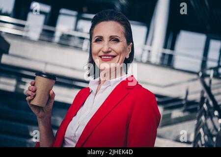 Foto di sicura signora assistente maturo indossare giacca rossa bere tè caldo fuori del complesso di affari della città Foto Stock