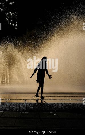 Una giovane ragazza, vista in silhouette, cammina su un marciapiede bagnato con le mani distese su un caldo tardo pomeriggio di agosto rinfrescato dallo spray dorato illuminato dal sole della fontana estiva a Karlsplatz, Monaco, Baviera, Germania. Foto Stock