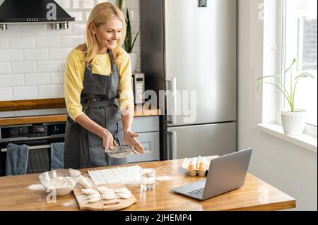 La donna bionda di mezza età cuoca a casa in cucina, casalinga in grembiule guarda una video ricetta su un computer portatile e lavora con l'impasto sul tavolo Foto Stock