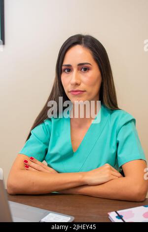 Una giovane infermiera femminile in uniforme verde con le braccia incrociate seduta davanti alla scrivania Foto Stock
