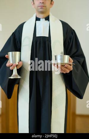 Clergyman in cassonetto con colletto clericale che tiene coppe con roba di comunione mentre pregava prima dell'oblazione e in attesa di parrocchiani Foto Stock