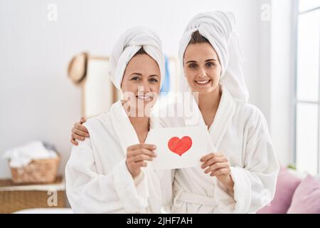 Donna e figlia di mezza età che indossano un accappatoio che tiene la carta del cuore sorridendo con un sorriso felice e fresco sul viso. Mostrando i denti. Foto Stock