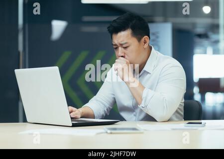 Uomo malato in ufficio tosse e ha naso che cola, uomo d'affari asiatico con freddo lavori in ufficio sul portatile Foto Stock