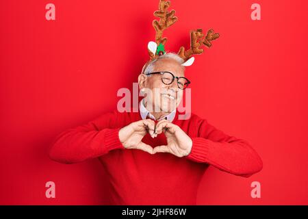 Uomo anziano con capelli grigi che indossa il cappello di natale del cervo sorridente nell'amore che mostra il simbolo del cuore e la forma con le mani. Concetto romantico. Foto Stock