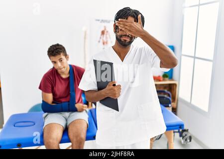 Giovane ispanico che lavora alla clinica di recupero del dolore con un uomo con il braccio rotto che copre gli occhi con la mano, guardando serio e triste. Senza vista, nascondendosi e. Foto Stock