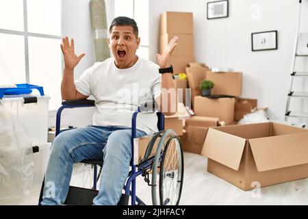 Giovane ispanico seduto su sedia a rotelle in una nuova casa che celebra la vittoria con un sorriso felice e l'espressione del vincitore con le mani sollevate Foto Stock