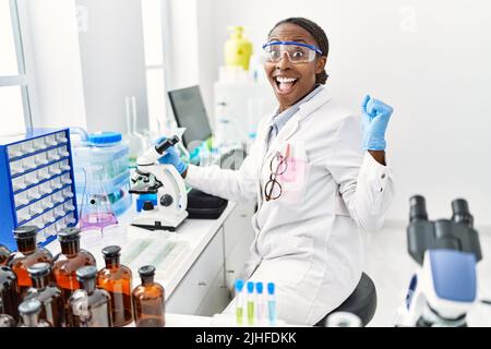 Donna africana che lavora al laboratorio di scienziato urlando orgoglioso, celebrando la vittoria e il successo molto eccitato con le braccia sollevate Foto Stock