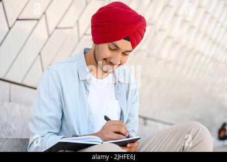 Focalizzato studente indiano in tradizionale turbano seduta all'aperto con notebook e prendere appunti, scrivere con penna, compilare diario, indù ragazzo che si prepara agli esami, rendendo programma seduta in paesaggio urbano Foto Stock