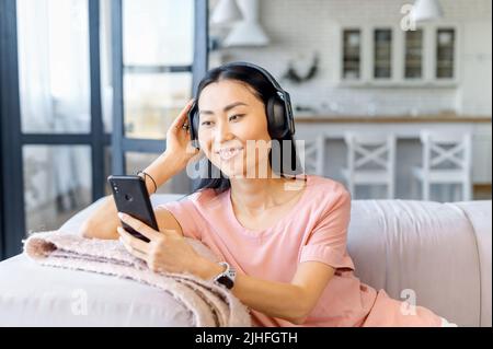 Donna asiatica attraente con capelli scuri e sorriso bianco neve, ascoltando la sua canzone preferita con le cuffie via telefono, seduto sul comodo divano, tenendo il braccio vicino alla testa, gioiosa, bella giornata Foto Stock