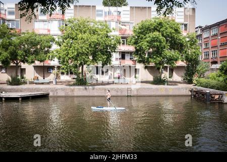 Un uomo su una tavola da paddle stand up vicino a un edificio di appartamenti su un canale in una giornata calda e soleggiata ad Amsterdam, Paesi Bassi. Foto Stock