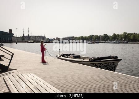 Una donna controlla il suo telefono mentre tiene la linea di ormeggio di un motoscafo su un canale in una giornata calda e soleggiata ad Amsterdam, Paesi Bassi. Foto Stock