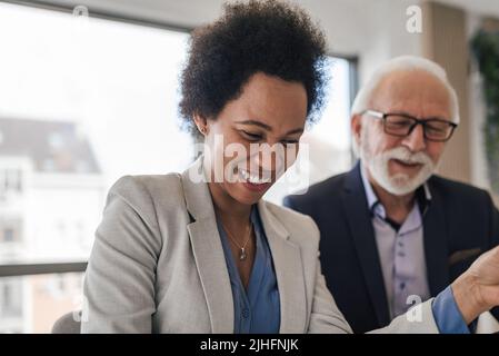 Donna multirazziale ridendo a scherzo divertente con diversi colleghi maschi senior in ufficio. Team di lavoro amichevole che guarda i documenti e gode di em positivo Foto Stock