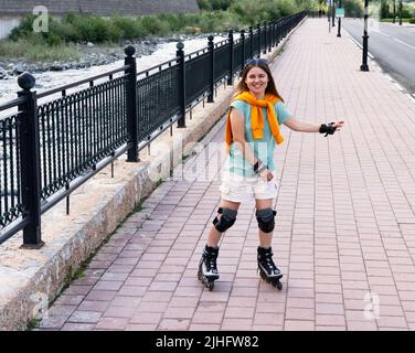 felice bella giovane donna in attrezzature di protezione che cavalcano su pattini a rotelle lungo il terrapieno in estate attività all'aperto, pattinaggio a rotelle Foto Stock