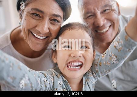 Ritratto di sorridenti nonni di razza mista e nipote prendendo un selfie nel salotto di casa. Uomo e donna ispanica senior che scatta foto e. Foto Stock