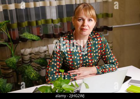 Defocus bionda donna che lavora al computer. Inizio o fine di una giornata lavorativa. Una giovane donna seduto al tavolo da lavoro e al computer portatile. Luogo di lavoro. Femmina. Verde Foto Stock