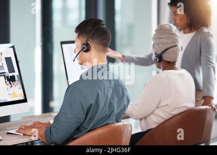 Agente di telemarketing ispanico del call center dal retro che parla con un auricolare mentre lavora con un computer in ufficio insieme ai suoi colleghi. Occupato Foto Stock