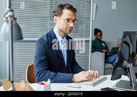 Uomo d'affari maturo in tuta digitando sulla tastiera seduta sul suo posto di lavoro di fronte al monitor del computer in ufficio Foto Stock