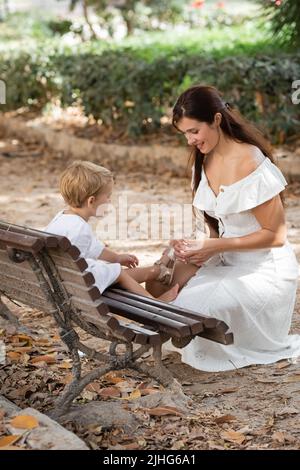 Donna sorridente che prende il sandalo dalla figlia del bambino in panchina nel parco di Valencia Foto Stock