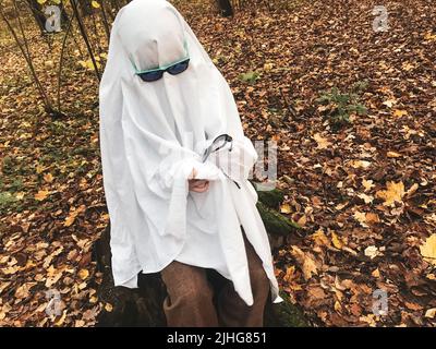 Un fantasma solitario nella foresta autunnale, in un foglio bianco con occhiali da sole, pulisce i vetri neri di un altro fantasma. Sfida fantasma Halloween. Foto Stock