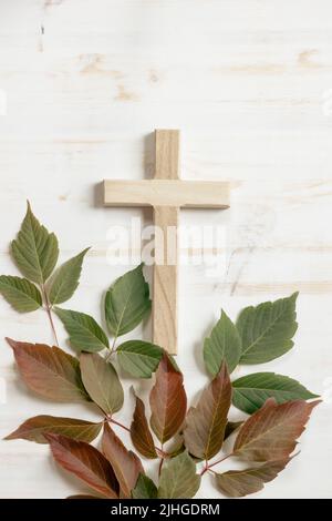 Semplice croce di legno cristiano su uno sfondo di legno bianco con un bordo di foglie autunnali mutevoli in verde e rosso con spazio copia Foto Stock