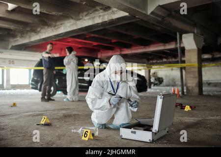 Giovane donna esperta di criminologia in indumenti da lavoro protettivi in cerca di impronte digitali di assassinio sulla scena del crimine contro due colleghi Foto Stock