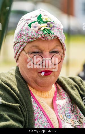 Ritratto della testa e della spalla di una donna anziana vestita con abbigliamento di classe operaia degli anni '40. Tipico scarafaggio con foulard, occhiali e fumatore Foto Stock