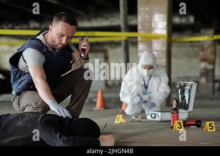 Ispettore detective o poliziotto in gilet bulletproof che tiene la lanterna piccola sopra il corpo morto mentre ispeziona la scena del crimine nell'area di parcheggio Foto Stock