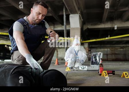 Ispettore detective in gilet bulletproof che tiene la lanterna piccola sopra il corpo morto mentre ispeziona la scena del crimine con esperto criminologico Foto Stock