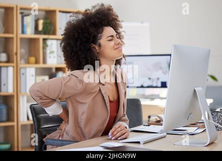 Una giovane donna di affari mista di corsa con un afro che soffre di mal di schiena mentre si siede alla sua scrivania in ufficio al lavoro. Un bellissimo africano Foto Stock
