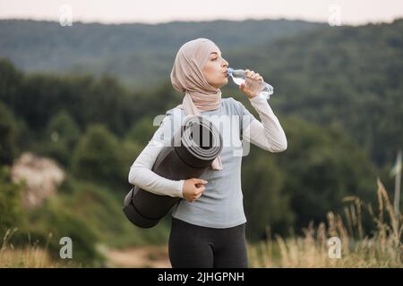 Vista laterale della bella giovane donna in hijab e activeWear acqua potabile da bottiglia mentre si trova in piedi al parco verde. Donna musulmana rilassante e rinfrescante dopo la pratica di yoga all'aperto. Foto Stock