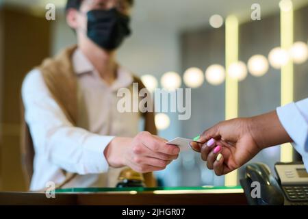 Mano di giovane donna nera che prende la keycard tenuta da ospite maschile in casualawear e maschera protettiva sul banco ricevimento Foto Stock