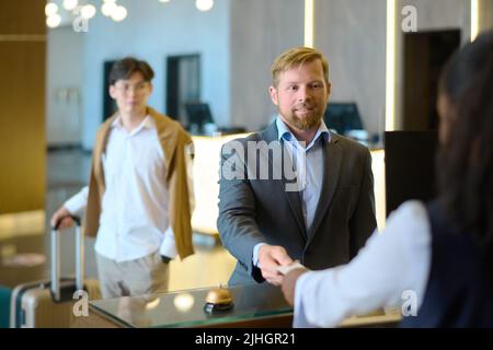 Un giovane uomo d'affari sorridente che ha dato la chiave di accesso all'addetta al ricevimento femminile al banco mentre lascia l'hotel per lavorare con un collega maschio Foto Stock